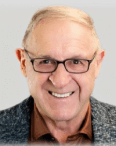 Smiling older man with glasses and light brown hair, wearing a dark blazer over a brown shirt, against a neutral background.