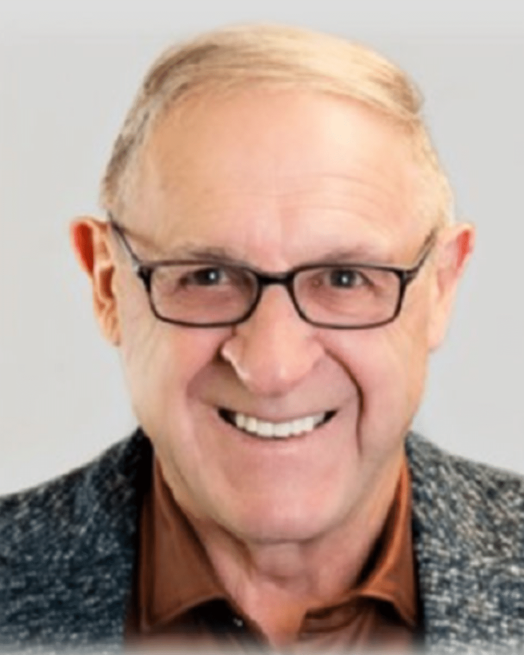 Smiling older man with glasses and light brown hair, wearing a dark blazer over a brown shirt, against a neutral background.