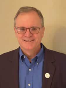 Smiling middle-aged man wearing glasses and a blue shirt under a dark blazer, standing against a neutral background.