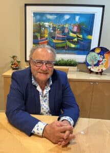 Smiling man with glasses seated at a wooden table, wearing a blue blazer over a floral shirt, with a colorful painting of boats in the background and decorative items on the table.