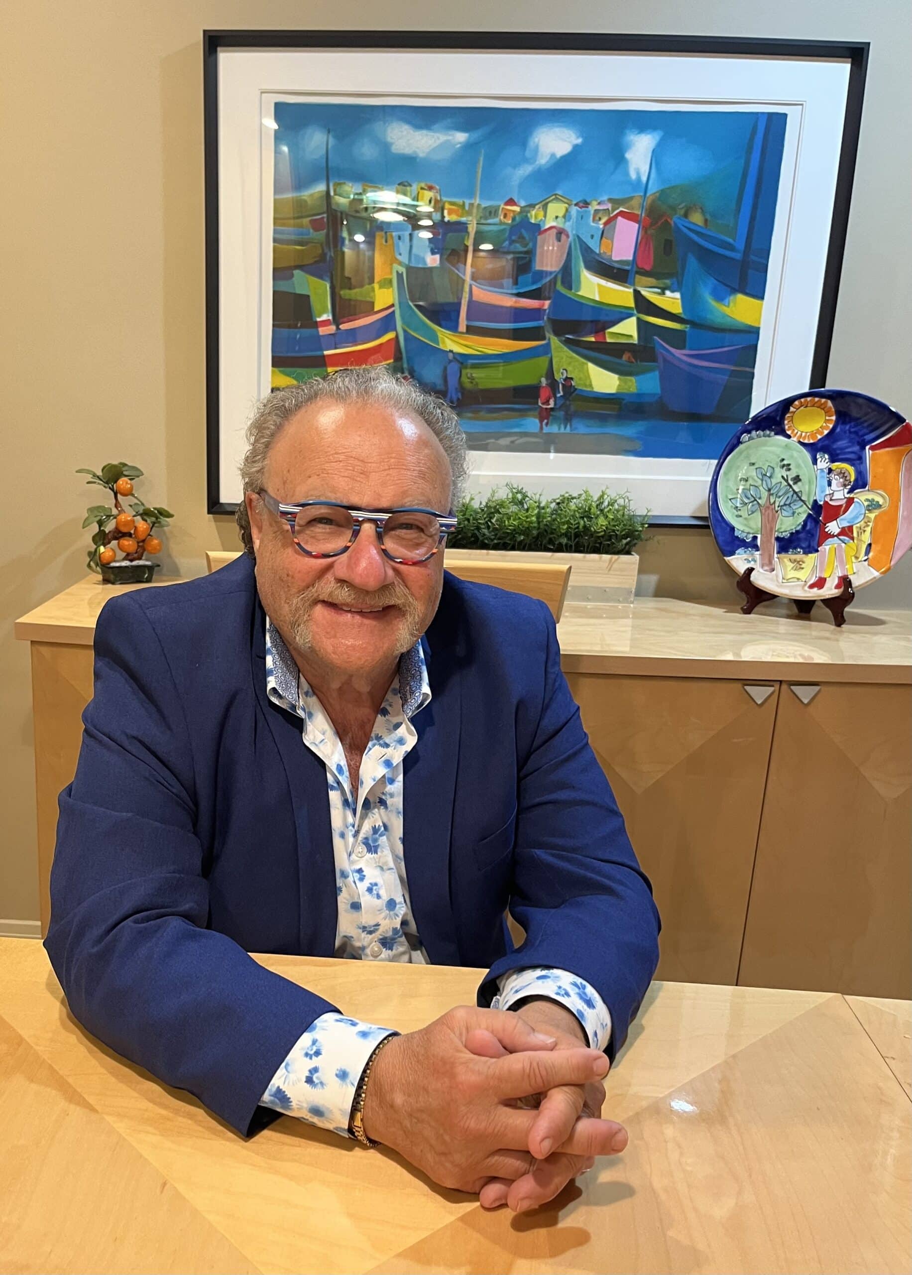 Smiling man with glasses seated at a wooden table, wearing a blue blazer over a floral shirt, with a colorful painting of boats in the background and decorative items on the table.